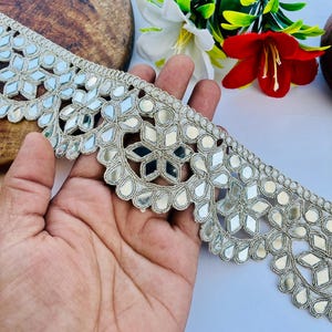 May include: Silver embroidered trim with mirrored accents. The trim features a repeating pattern of geometric shapes and floral designs. The trim is held in a hand, with a wooden surface and flowers in the background.