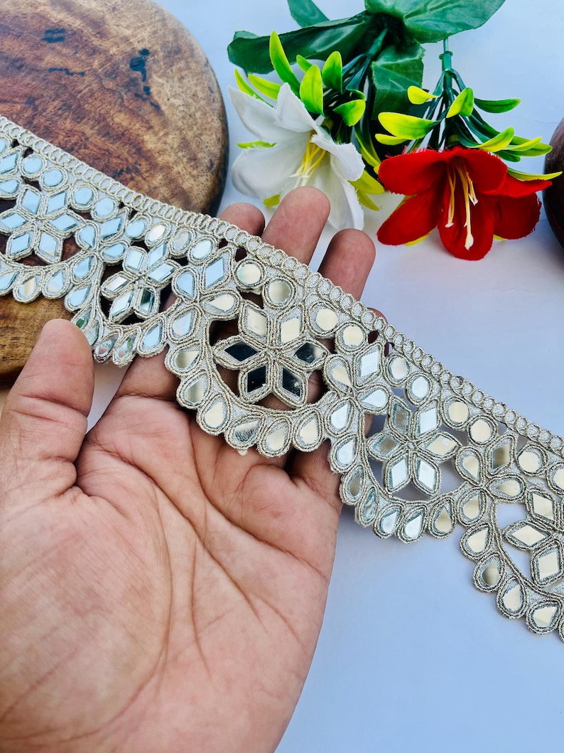 May include: Silver embroidered trim with mirrored accents. The trim features a repeating pattern of geometric shapes and floral designs. The trim is held in a hand, with a wooden surface and flowers in the background.