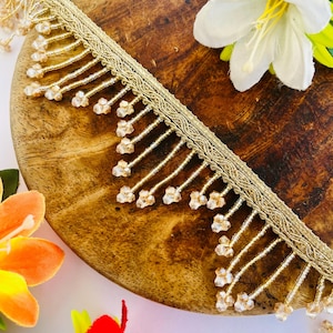 Puede incluir: Ribete dorado con cuentas con un diseño delicado. El ribete presenta una base tejida con hebras colgantes de pequeñas cuentas transparentes. El ribete se muestra sobre una superficie de madera con flores coloridas en el fondo. Este ribete es adecuado para varios proyectos de manualidades.