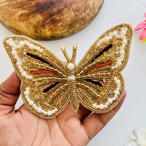 Può includere: Un ornamento a forma di farfalla color oro, riccamente decorato con perline dorate, bianche e arancioni. Le ali sono bordate con piccole perline bianche e presentano un motivo dettagliato. La farfalla è tenuta in una mano.