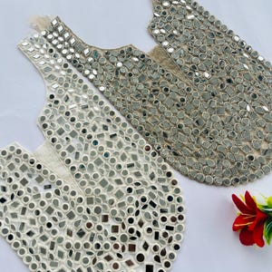 May include: Two decorative silver mirror embroidered fabric pieces. The pieces are in a vest shape, with a cream-colored backing and a variety of small, reflective mirrors in different shapes. A red flower with green leaves is in the background.