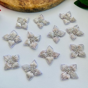 Può includere: Una collezione di applicazioni floreali ricamate color argento. Ogni applicazione presenta quattro petali con un design strutturato e a coste e un piccolo centro dettagliato. Le applicazioni sono sparse su una superficie bianca, con uno sfondo in legno.