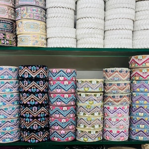 May include: Multiple stacks of decorative fabric ribbon trim arranged on shelves. The ribbons feature intricate embroidered floral and geometric patterns in various colors including black, white, blue, pink, yellow, and light gray, organized in rolls.