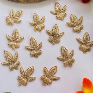 May include: Gold-colored leaf-shaped embroidered appliques. The appliques are arranged on a white surface. Each leaf has detailed stitching, creating a textured appearance. These decorative appliques are suitable for various craft projects.