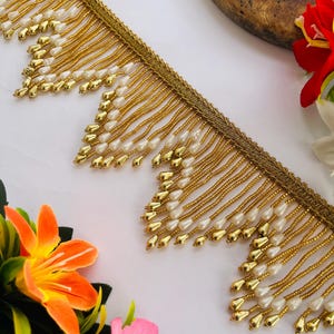 May include: Gold beaded fringe trim with white teardrop-shaped beads. The trim has a woven gold border and is arranged on a white surface with colourful flowers in the background. This decorative trim is suitable for various craft projects.
