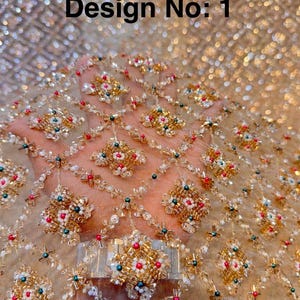 May include: A sheer gold fabric featuring a diamond-shaped grid pattern embellished with gold, white, and red beads, along with small green accents. The textile displays the text Design No: 1 in black lettering across the upper portion of the image.