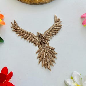 Puede incluir: Un parche bordado de pájaro en color dorado con un intrincado trabajo de hilo metálico. El pájaro se muestra con las alas completamente extendidas en vuelo, exhibiendo patrones de plumas detallados en las alas y la cola con textura.