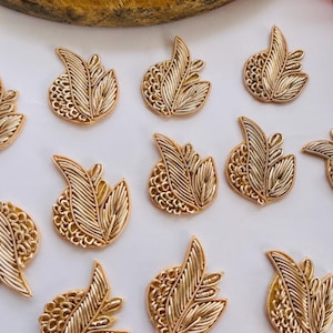 May include: Gold-colored leaf-shaped embellishments with intricate wire detailing. The leaves have a textured, layered design, with a mix of solid and openwork patterns. The embellishments are arranged on a white surface.