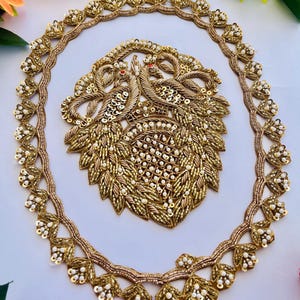 May include: A gold embroidered neckline applique featuring a pair of peacocks and a central floral motif, accented with white beads and sequins. The set includes a matching oval-shaped border trim decorated with repeating leaf-like patterns.