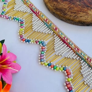 Puede incluir: Ribete decorativo con borde festoneado. El ribete presenta cuentas blancas, varillas doradas y cuentas coloridas en forma de lágrima. El borde superior tiene una cinta estampada con rayas rosas, verdes y amarillas. El ribete está sobre una superficie blanca.