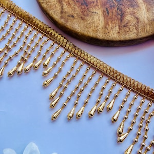 May include: A gold-coloured decorative trim featuring a woven fabric header and a series of dangling fringe strands. Each strand consists of small round beads leading to a larger, teardrop-shaped pendant, all finished in a metallic gold tone.