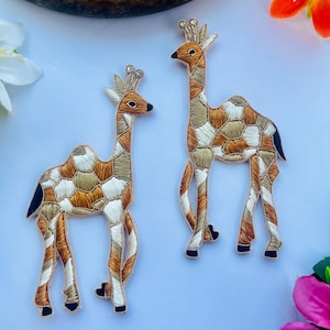 May include: Two embroidered patches shaped like giraffes with camel-like humps. The patches feature a mosaic pattern of white, tan, and copper-colored thread with gold metallic outlines. Each giraffe has a small crest with clear rhinestones on its head.