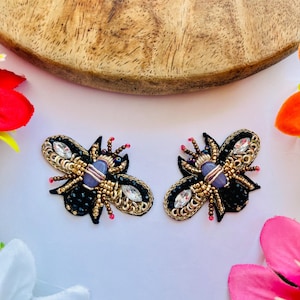 May include: Two black and gold beaded bee-shaped embellishments with clear and purple accents. The wings are black with gold trim, and the body features a purple gem. These decorative pieces are set against a white background.