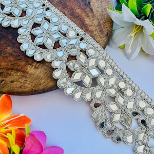 May include: Silver embroidered trim with mirrored accents. The trim features a repeating floral and geometric pattern. The trim is displayed on a wooden surface with colorful flowers in the background. This trim is suitable for various craft projects.