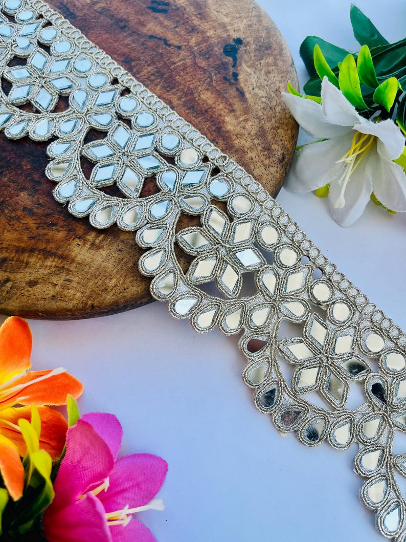 May include: Silver embroidered trim with mirrored accents. The trim features a repeating floral and geometric pattern. The trim is displayed on a wooden surface with colorful flowers in the background. This trim is suitable for various craft projects.