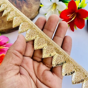 May include: Gold sequin trim with a scalloped edge. The trim features a leaf design and is held in a hand. White and red flowers are in the background.