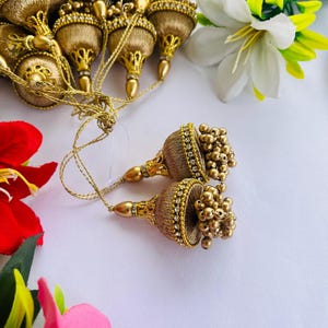 Puede incluir: Borlas doradas con intrincados abalorios y un diseño en forma de lágrima. Las borlas están dispuestas con acentos florales, incluyendo lirios blancos y amarillos y flores rojas y rosas, sobre un fondo blanco. Las borlas son probablemente para fines decorativos.