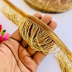 Puede incluir: Un fleco de cuentas doradas que presenta una banda de tela tejida con múltiples filas de cuentas de vidrio metálicas doradas colgantes. Este borde decorativo está diseñado para proyectos de costura y adornos.