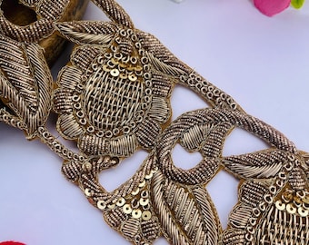 May include: Close-up of a gold embroidered trim with a floral and leaf design. The trim features intricate detailing with sequins and metallic threads. The design includes a repeating pattern of flowers and leaves.
