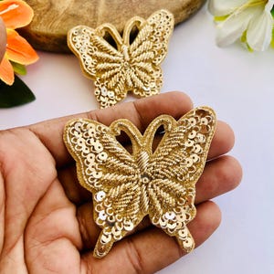 Puede incluir: Dos parches dorados con forma de mariposa que presentan bordados de hilo metálico y pequeñas lentejuelas circulares en los bordes de las alas. Los parches tienen una apariencia texturizada con patrones de costura en relieve y diseño simétrico.