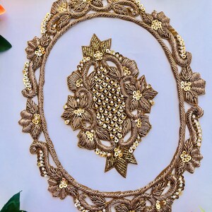 May include: An oval-shaped gold embroidered applique featuring a central lattice pattern and floral motifs. The design is embellished with metallic gold thread, small gold sequins, and tiny white beads, creating a textured and ornate decorative piece.