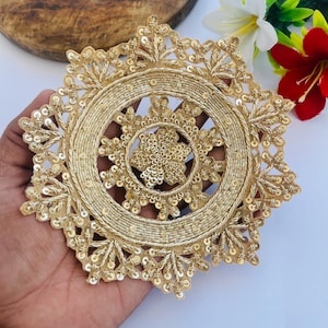 Op de afbeelding: Goudkleurige, cirkelvormige geborduurde applicatie met ingewikkelde details. De applicatie heeft een centraal bloemenontwerp, een kralenring en een geschulpte buitenrand. Het ontwerp is versierd met pailletten, wat een glinsterend effect creëert.