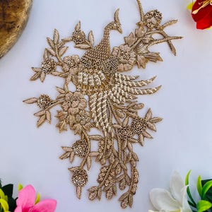 Op de afbeelding: Goudkleurige geborduurde pauwenapplicatie met bloemen- en blad details. De applicatie heeft een gedetailleerd ontwerp met een pauw in het midden, omringd door bloemen en bladeren. Perfect voor het verfraaien van kleding of knutselwerk.