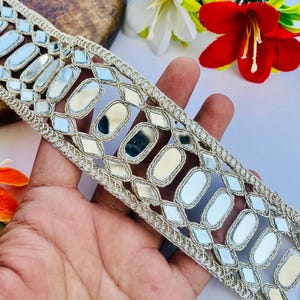 Puede incluir: Un adorno decorativo con un patrón repetido de rectángulos y diamantes espejados, delineados con hilo dorado. El adorno se sostiene en una mano, con flores blancas y rojas en el fondo. Adecuado para embellecer prendas o manualidades.