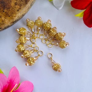 May include: Gold-coloured beaded ornaments with a spherical design, each attached to a small gold ring. The beads have a textured appearance and are arranged in a semi-circle on a white surface, with pink and white flowers in the background.