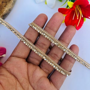 May include: Gold-colored decorative trim with clear, sparkling rhinestones. The trim is held in a hand, with a white and red flower in the background. The trim is likely used for embellishing clothing or crafts.