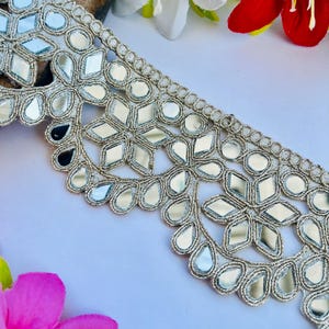 May include: Silver embroidered trim with mirrored accents. The trim features a floral and geometric design with reflective mirror pieces. The trim is on a white surface with red, white, and pink flowers in the background.