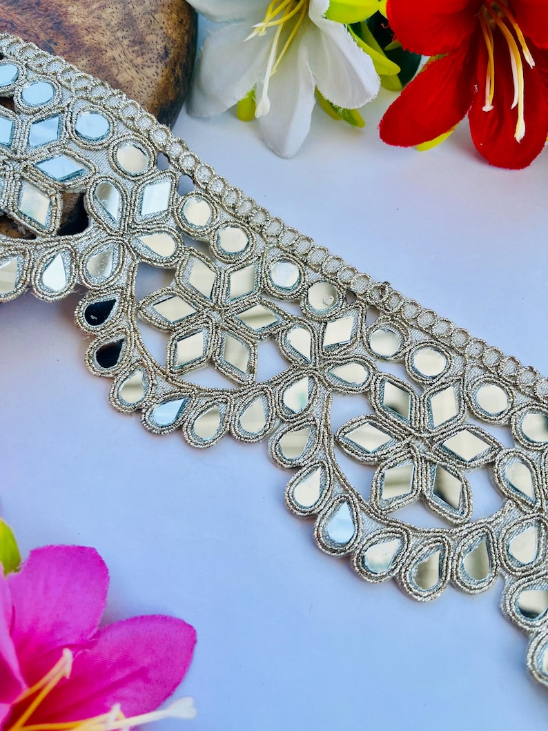 May include: Silver embroidered trim with mirrored accents. The trim features a floral and geometric design with reflective mirror pieces. The trim is on a white surface with red, white, and pink flowers in the background.