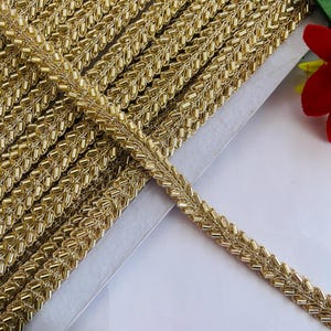 May include: Close-up of a roll of gold beaded trim. The trim has a woven design with small, gold beads. The trim is on a white surface, with a few colorful flowers in the background. This trim is suitable for sewing and craft projects.