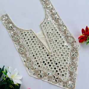 Può includere: Un ornamento per abito bianco con scollatura a V con intricati ricami a specchio e floreali. Il design presenta piccoli specchi rotondi e delicati fili color oro, creando un aspetto decorativo e ornato. I bordi sono rifiniti con un fine bordo bianco.