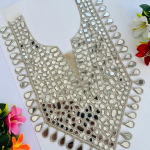 May include: A silver-coloured, decorative neckline with a V-neck design. The neckline is embellished with numerous small, reflective mirror pieces in various shapes. The edges are trimmed with silver thread. Ideal for embellishing clothing.