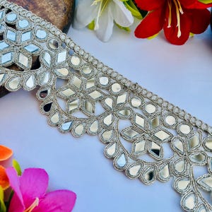 May include: Silver mirror embroidered trim with a scalloped edge. The trim features a repeating pattern of mirrored shapes and teardrop designs, with a metallic silver thread. Decorative trim for crafts and sewing.