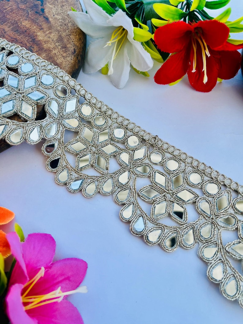 May include: Silver mirror embroidered trim with a scalloped edge. The trim features a repeating pattern of mirrored shapes and teardrop designs, with a metallic silver thread. Decorative trim for crafts and sewing.
