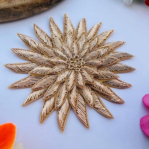 Peut inclure: Une applique florale brodée de couleur or, avec un design détaillé et superposé. L'applique présente une fleur centrale avec des pétales en forme de feuilles rayonnantes. La broderie complexe crée un effet texturé en trois dimensions, adapté à l'embellissement de vêtements ou d'objets artisanaux.