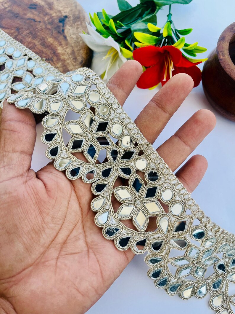 May include: A decorative trim with a mirrored design. The trim features a repeating pattern of mirrored shapes, including diamonds and circles, set against a gold-colored fabric. The trim is likely used for embellishing clothing or crafts.