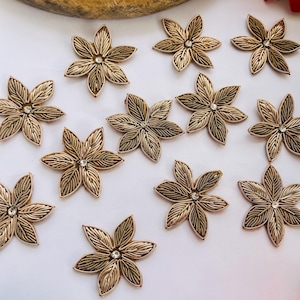 Pode incluir: Enfeites florais dourados com um strass central. As flores têm desenhos detalhados de pétalas e estão dispostas sobre uma superfície branca. Estes elementos decorativos podem ser usados para artesanato ou moda.