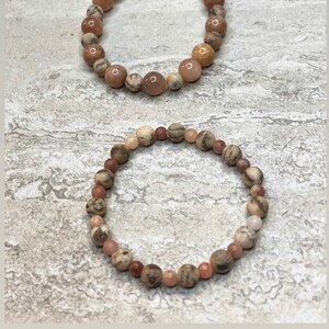 May include: Two beaded bracelets made with sunstone, sesame jasper, and feldspar. The bracelets are brown, pink, and white.