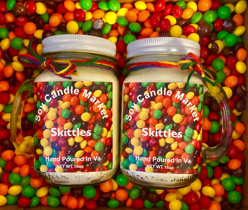 Op de afbeelding: Twee glazen potten met deksels, gevuld met een witte kaars. De potten hebben een kleurrijk etiket met de tekst "Soy Candle Market" en "Skittles". De potten zijn omringd door kleurrijke snoepjes.