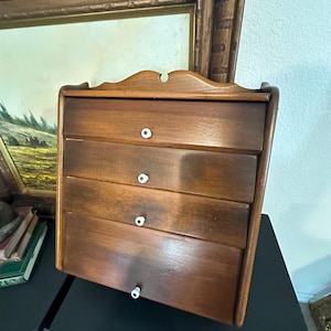 May include: A wooden wall-mounted storage unit with four drawers and white knob pulls. The drawers are made of a light brown wood with a smooth finish.