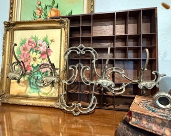 Può includere: Un attaccapanni da parete antico in ottone con volute ornate e tre ganci. L'attaccapanni è esposto con dipinti floreali incorniciati e una mensola a cubi in legno vintage. Sono visibili anche una pila di vecchi libri e un piccolo oggetto di metallo.
