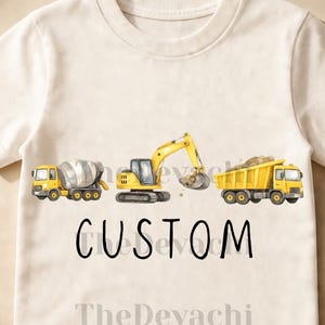 May include: Cream-colored t-shirt featuring a watercolor illustration of construction vehicles: a cement mixer, excavator, and dump truck. The word "CUSTOM" is printed in black script below the vehicles, with "TheDevachi" below that.