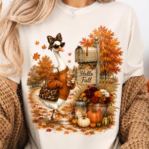 May include: A cream-colored t-shirt featuring a graphic of a goose wearing a leopard print bow, sunglasses, and an orange sweater while holding a coffee cup. The scene includes a mailbox with the text Hello Fall, autumn leaves, and pumpkins.