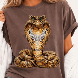 Puede incluir: Una camiseta marrón de manga corta con el gráfico de una cobra dorada que lleva gafas con estampado de leopardo y un collar plateado estilo diamante. La serpiente tiene ojos verdes y está enroscada en la base, mostrando escamas en tonos dorados.