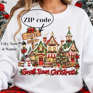 May include: A white sweatshirt featuring a colorful graphic of a snowy village scene with a snowman, a decorated Christmas tree, and houses. The graphic includes a wooden sign labeled 05672 and STOWE VT, with the text Small Town Christmas below.