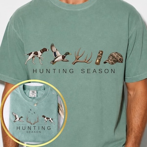 May include: A sage green t-shirt featuring a graphic print with the text HUNTING SEASON. The design includes illustrations of a spotted hunting dog, a flying mallard duck, deer antlers, a shotgun shell, and a camouflage baseball cap.