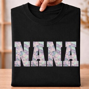 Może przedstawiać: Czarny t-shirt z napisem "NANA" w dużych literach w stylu patchwork na przodzie. Litery wypełnione są pastelowym wzorem pikowanym w odcieniach fioletu, błękitu i zieleni. Koszulka jest złożona i trzymana przez dłoń.